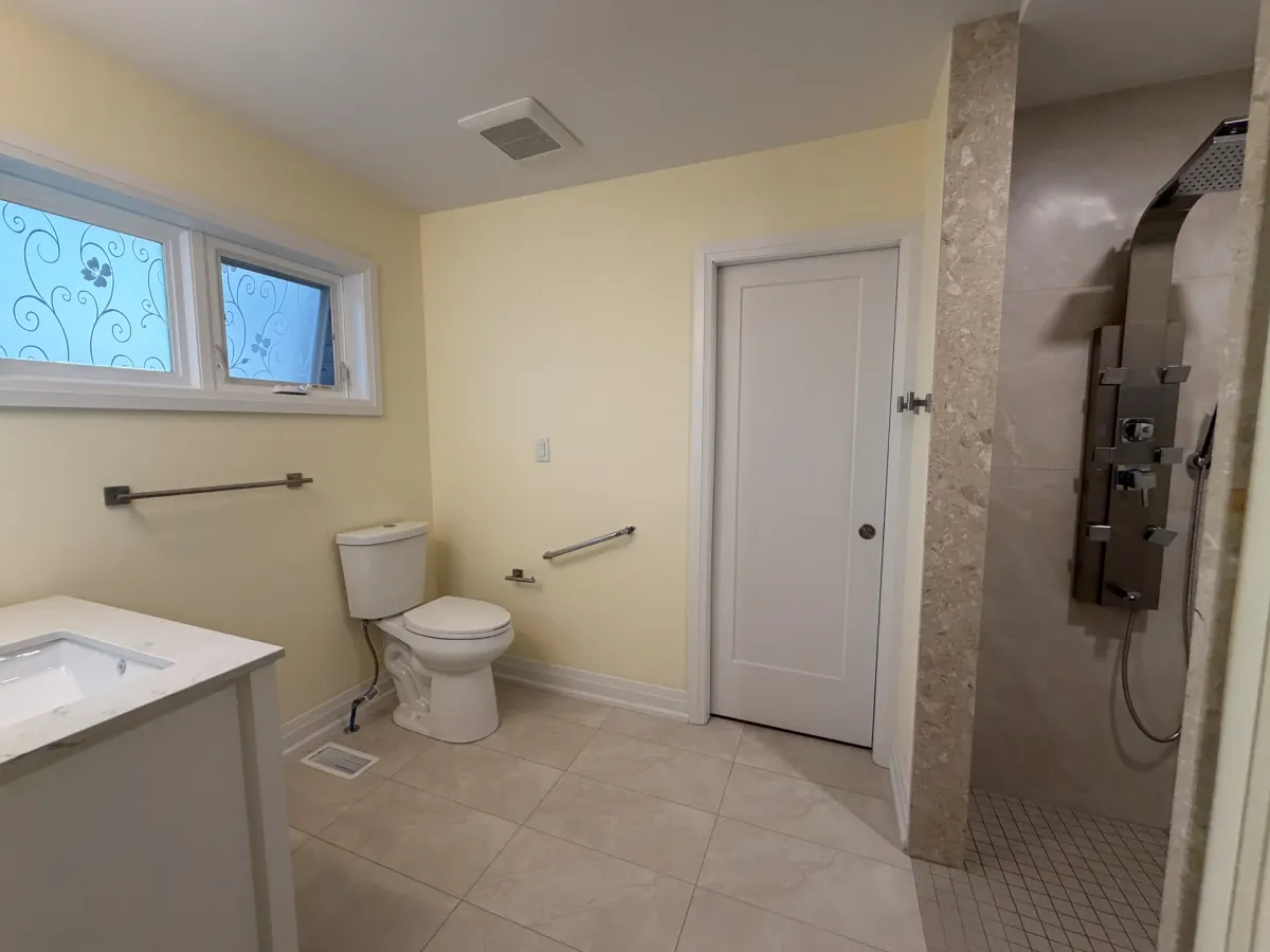 After: the open room with a curbless walk-in shower, a grab bar by the toilet, a white vanity and the frosted window