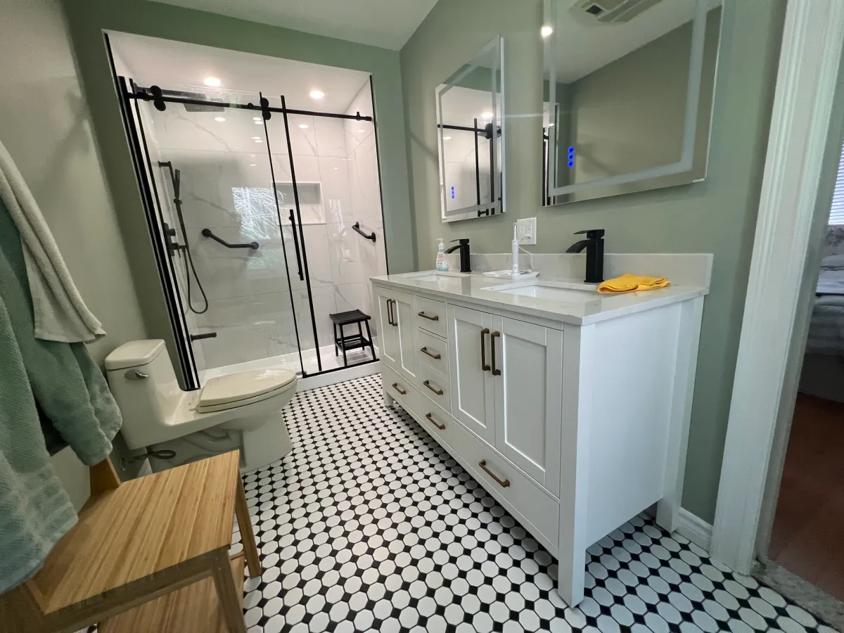 After: a black-framed sliding glass shower with a grab bar, a white shaker vanity with a quartz top and black fixtures, on a black-and-white mosaic floor with sage green walls