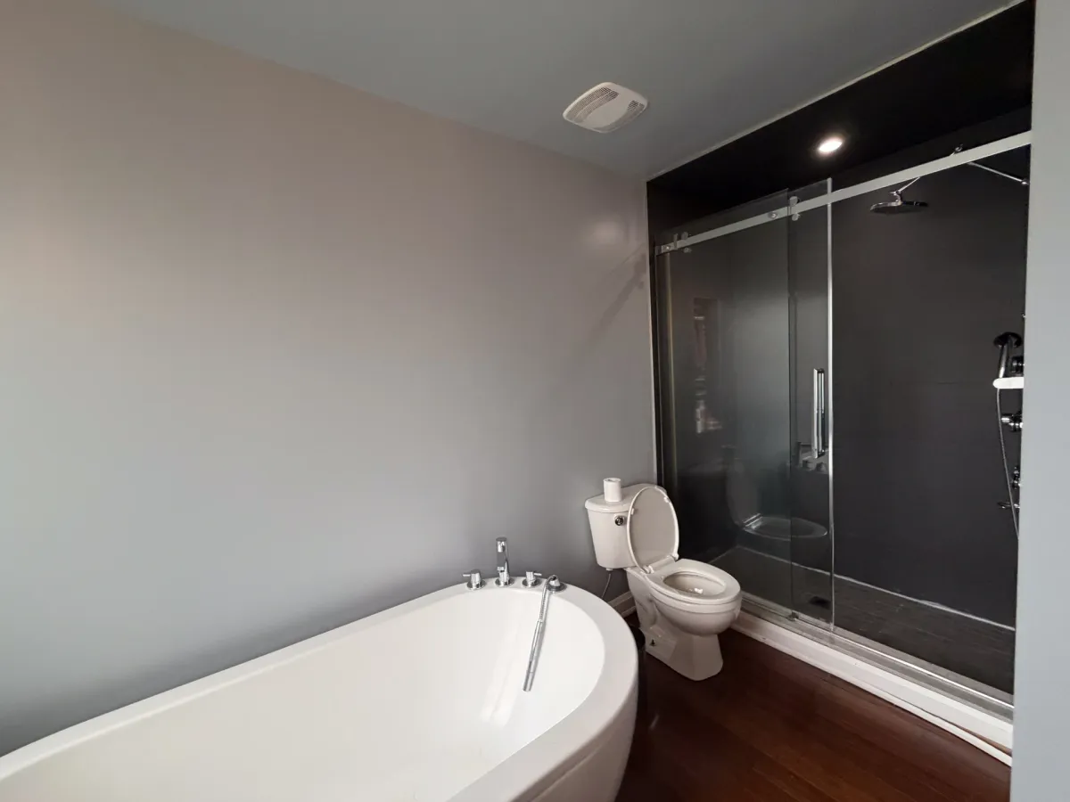 Before: a freestanding tub and the old toilet beside a dark-tinted sliding glass shower, on cherry hardwood