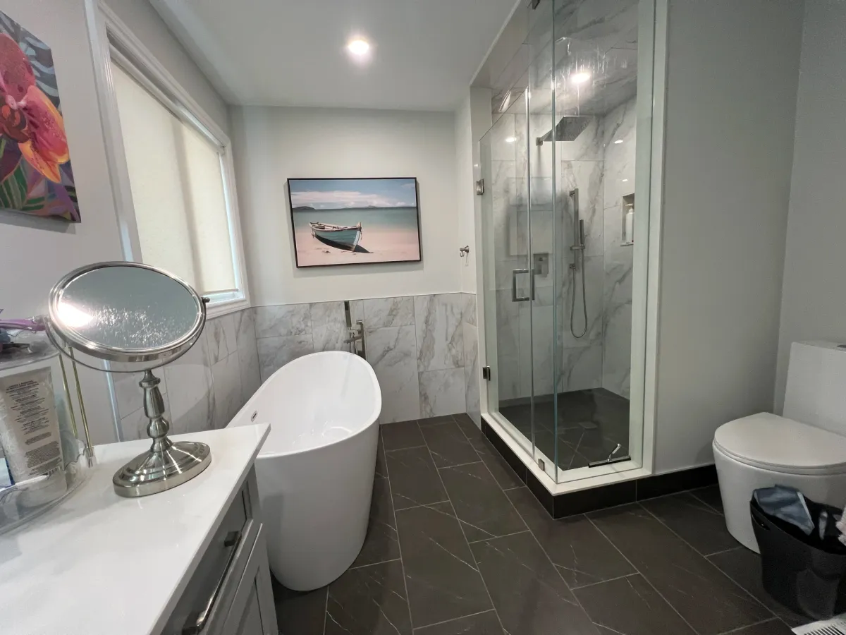 After: a freestanding tub under the window, a frameless glass shower with a marble-look surround and a grey vanity on dark porcelain tile