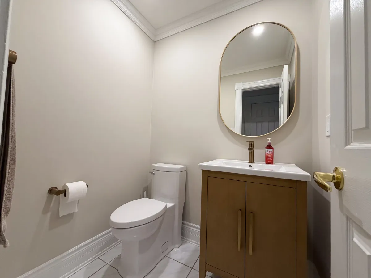After: an oak vanity with a brushed-gold faucet under an oval brass-framed mirror, a new toilet and off-white walls