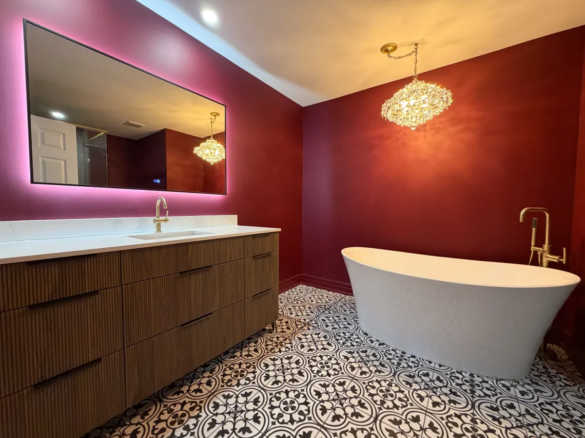 After: a fluted walnut vanity, a freestanding tub with a brass floor-mounted filler and a crystal chandelier, on black-and-white patterned tile against burgundy walls