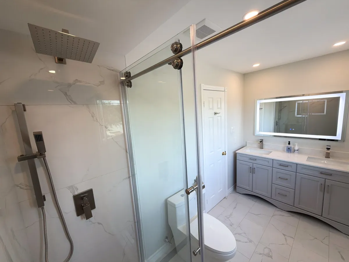 After: a marble-look walk-in shower with a rain head behind sliding glass, and a grey double vanity under one wide backlit mirror