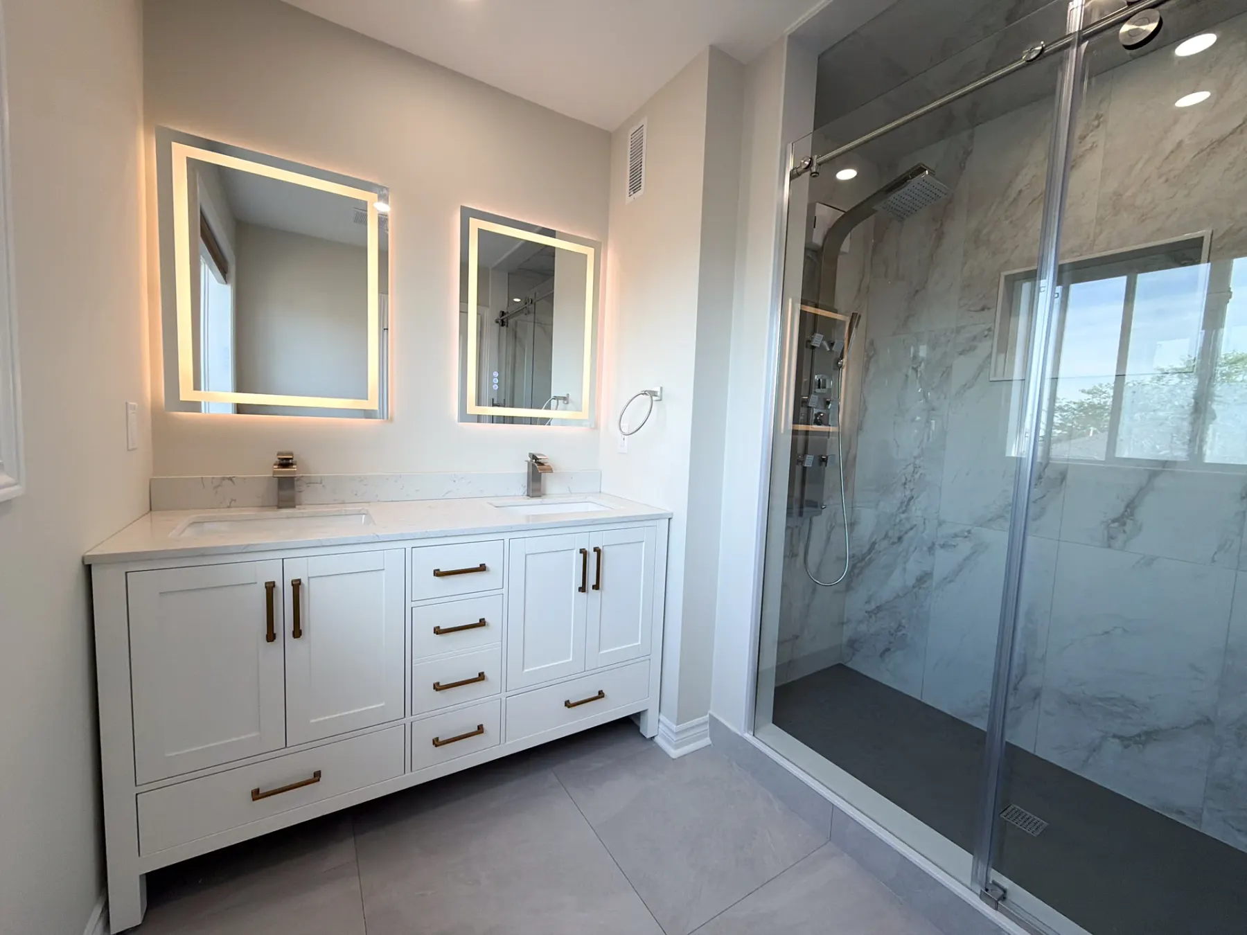 Four-piece bathroom with a white double vanity, two backlit mirrors and a marble-look tiled shower behind sliding glass