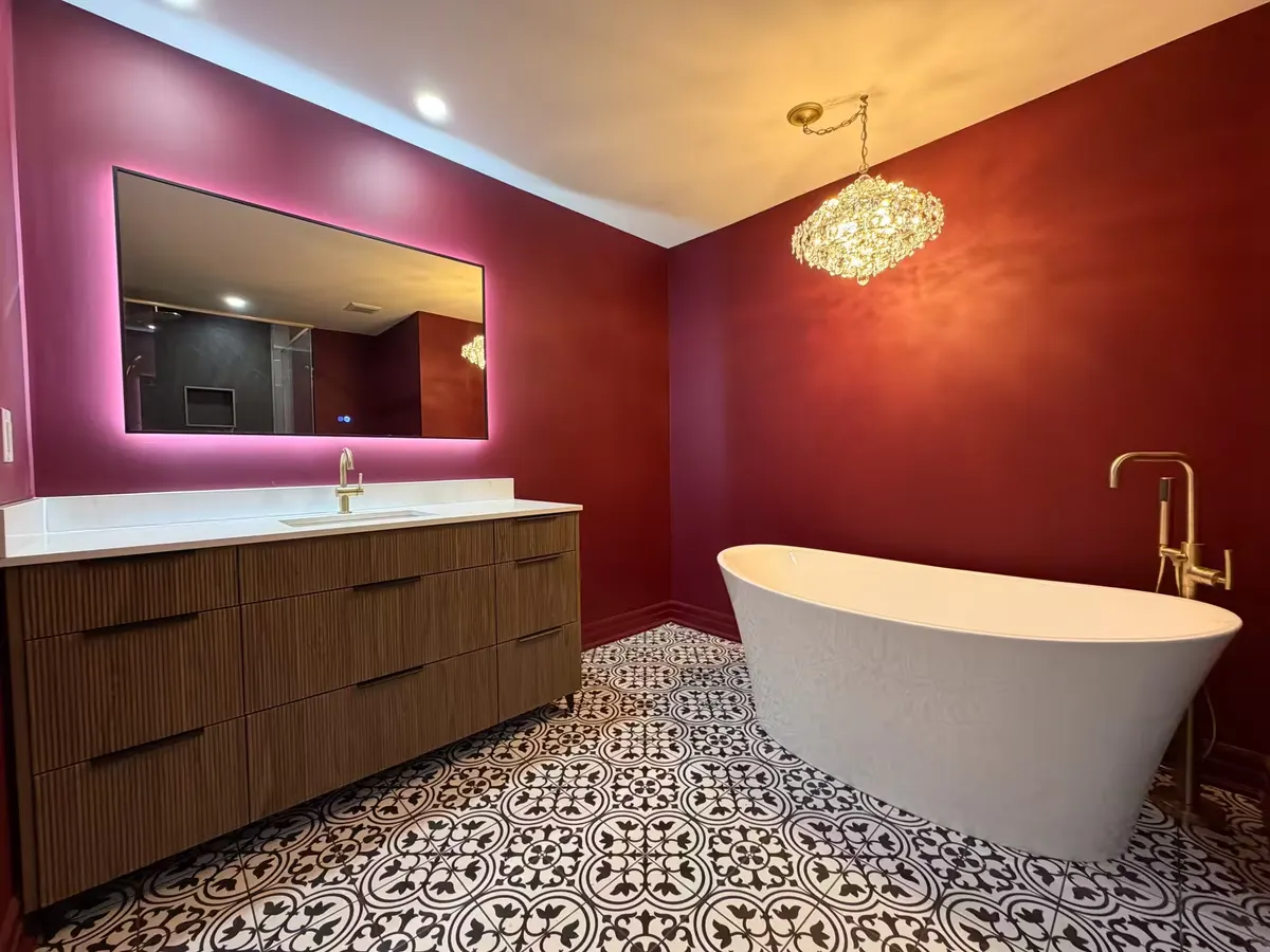 Ensuite with deep red walls, a freestanding white tub, a brass floor-mounted filler, a fluted walnut vanity under a backlit mirror and a crystal chandelier, on black-and-white patterned tile