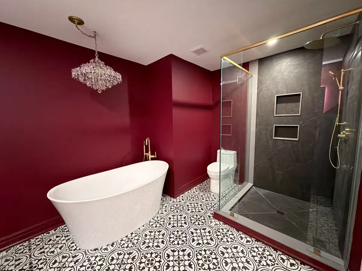 Freestanding white tub beside a dark slate-look walk-in shower trimmed in brass, under a crystal chandelier on black-and-white patterned tile