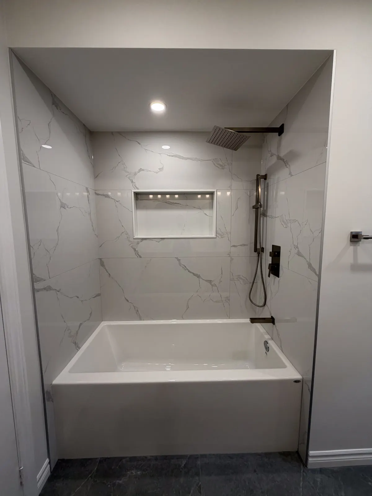 Alcove tub with a marble-look tile surround, a lit storage niche and a brushed-brass rain shower