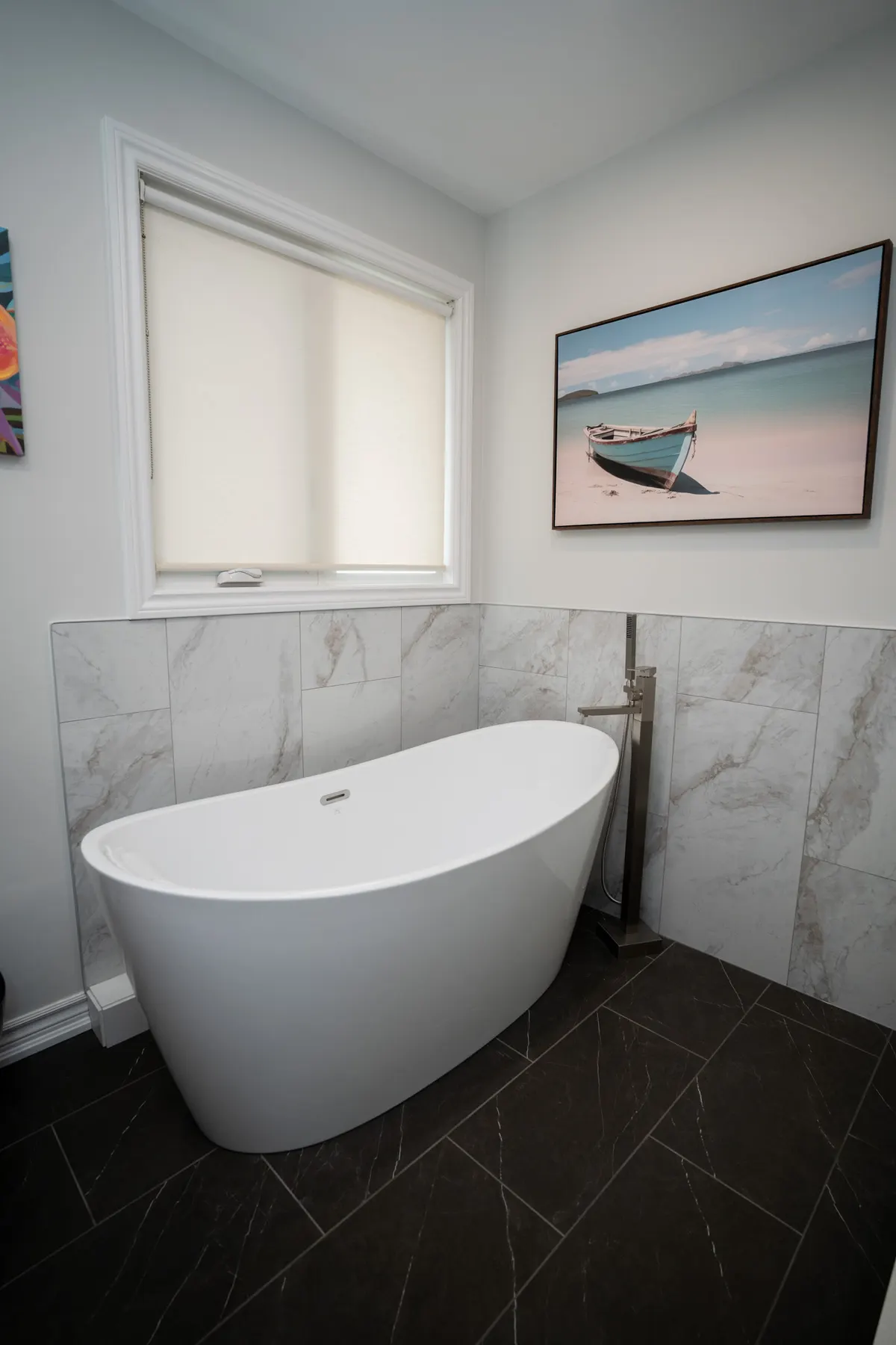 Freestanding soaker tub with a floor-mounted filler against marble-look half-height tile, on a dark slate-look floor