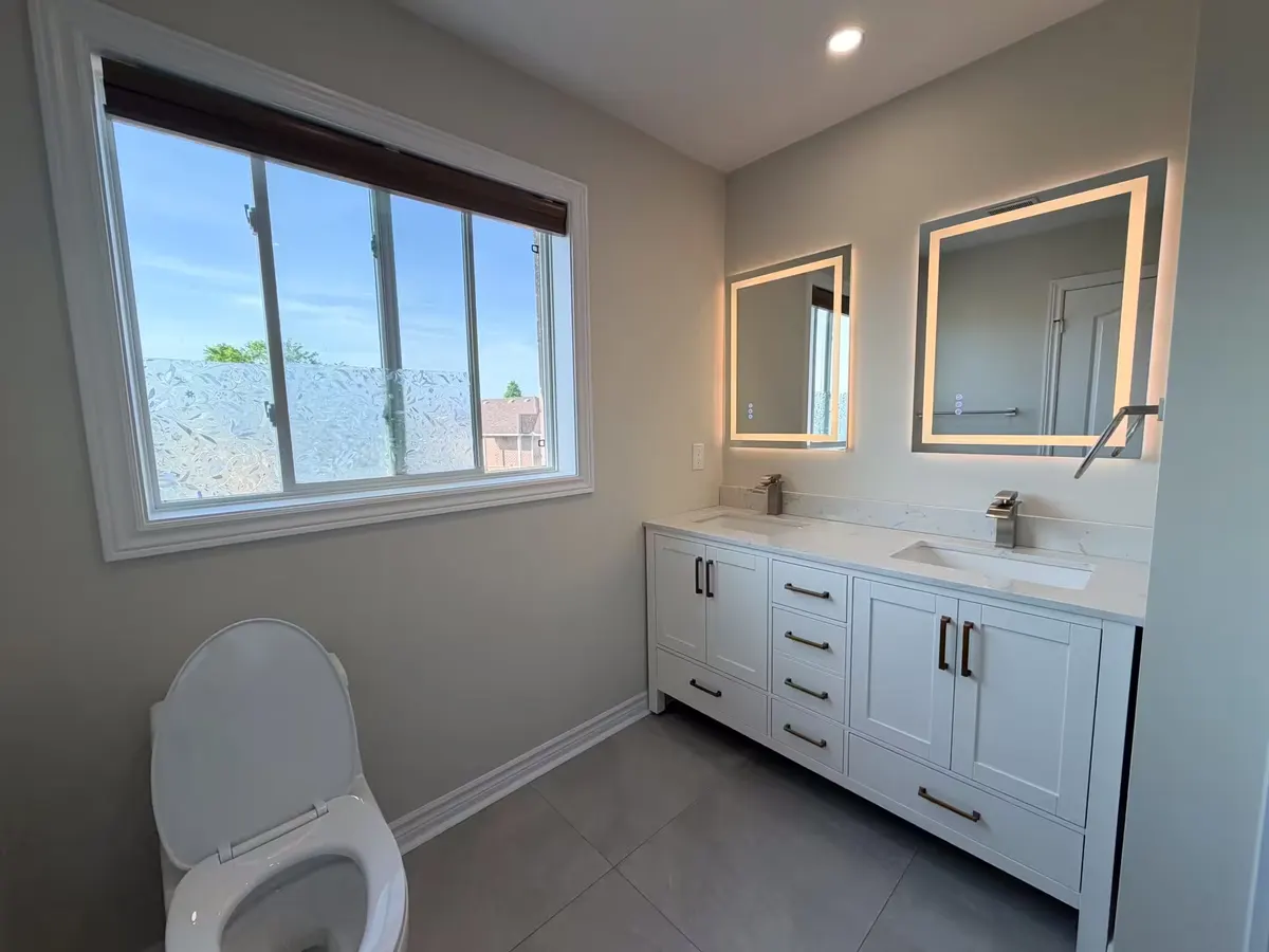 Bathroom with a white shaker double vanity, two backlit mirrors, grey large-format floor tile and a wide window