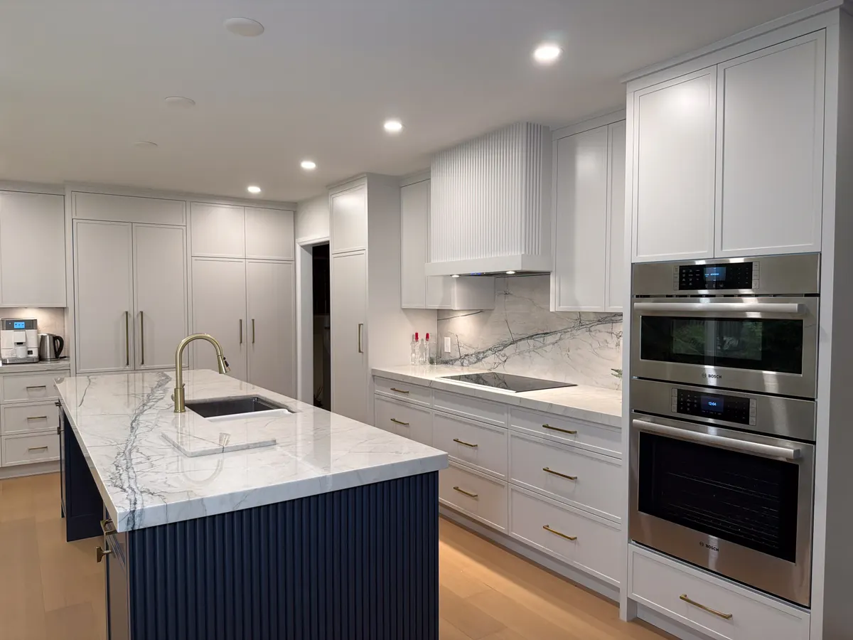 White kitchen with a navy island, built-in wall ovens and marble counters